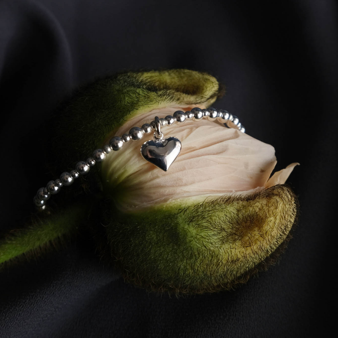 Photograph of a silver bracelet by Annie Haak, sitting on top of a pink poppy. The bracelet has a single silver heart-shaped charm