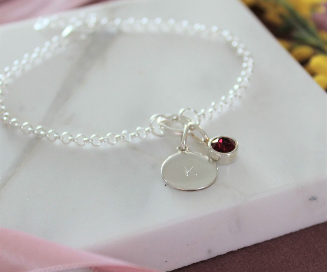 Photograph of a silver bracelet with a birthstone charm with a red gem and a silver disc personalised with the initial 'K' engraved on it
