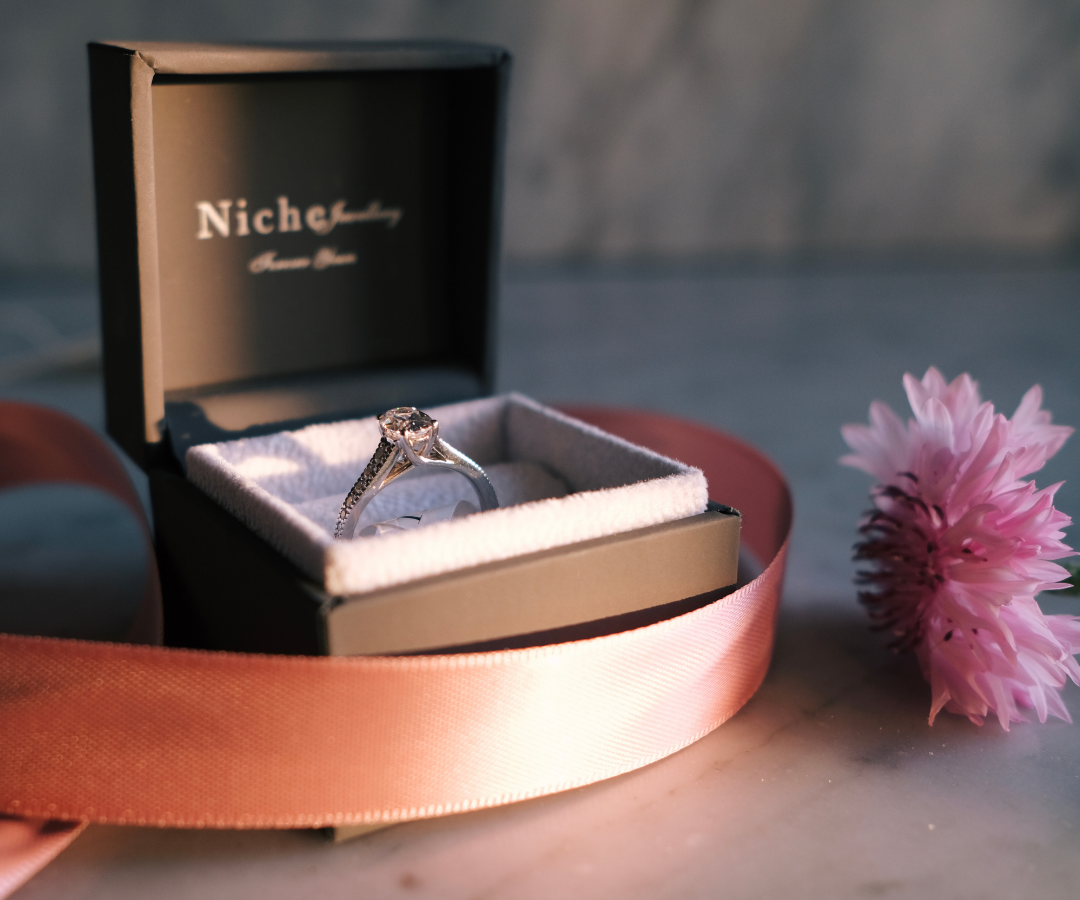 Photograph of an engagement ring in a box with a pink ribbon and flower beside it