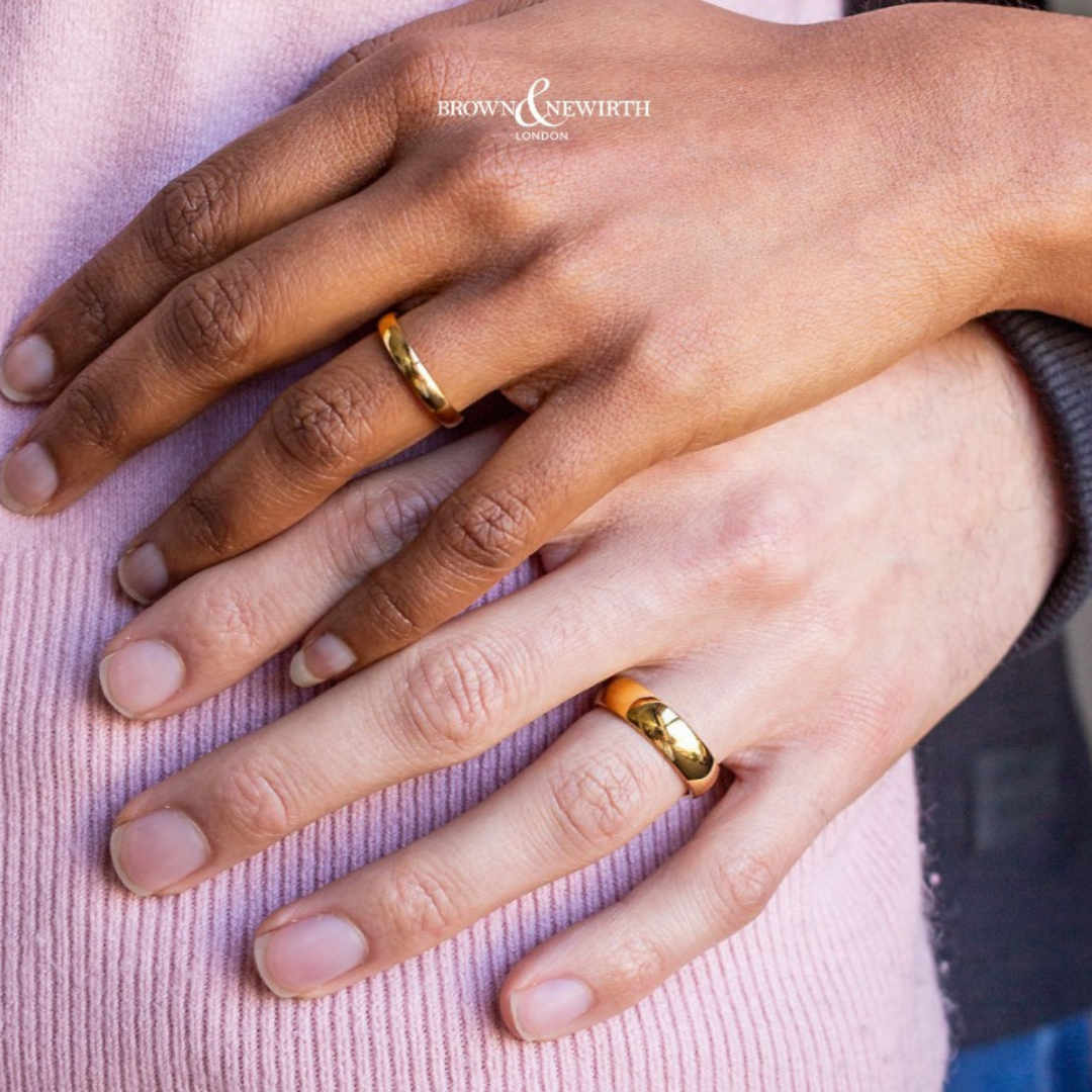 Photograph shows two hands each wearing a gold wedding band by brown & newirth
